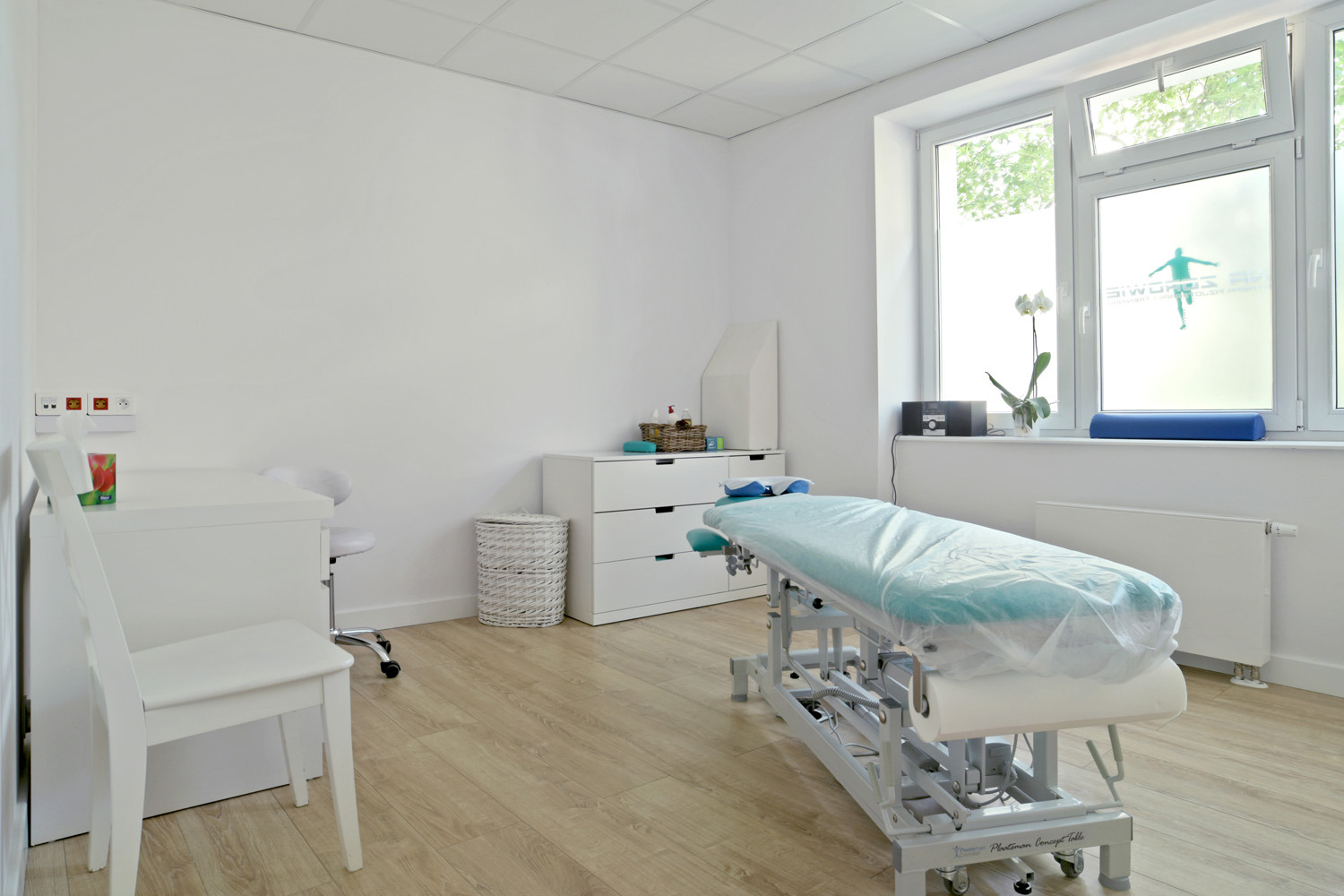Jasne, minimalistyczne wnętrze gabinetu z łóżkiem rehabilitacyjnym przykrytym folią ochronną, biurkiem, krzesłem i komodą. Na oknie widoczna naklejka z sylwetką ćwiczącej osoby.