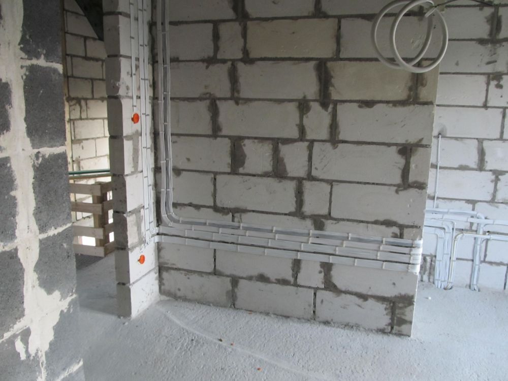 Surowe wnętrze w trakcie prac wykończeniowych: ściany z białych bloczków, widoczne kable elektryczne w białych listwach przypodłogowych i peszlach, instalacja prowadzona po ścianie, widoczne otwory...