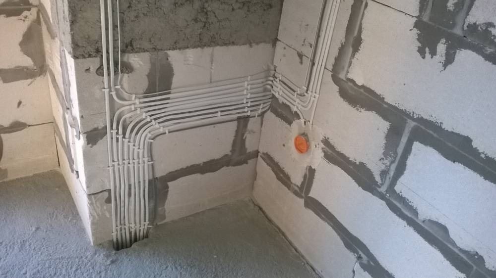 Białe kable elektryczne układane natynkowo w rogu surowego pomieszczenia z bloków betonowych, widoczne mocowania kabli i puszka instalacyjna.