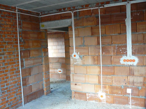 Surowy mur z cegły dziurawki z widoczną instalacją elektryczną: białe kable prowadzone po ścianach i w peszlach, puszki podtynkowe z pomarańczowymi dekielkami. Budynek w stanie deweloperskim.