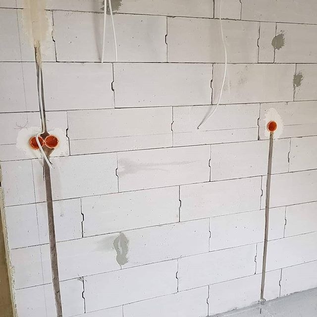 Biała ściana z betonu komórkowego z wyżłobionymi bruzdami pionowymi, w których poprowadzone są kable elektryczne i zamontowane puszki instalacyjne w kolorze pomarańczowym.