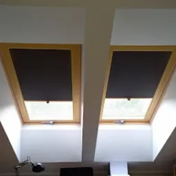 Dwa okna dachowe z zamontowanymi, zwiniętymi roletami w kolorze ciemnoszarym, drewniane ramy okienne, widoczne fragmenty otoczenia za oknami, białe wnęki okienne, lampka biurkowa w dolnej części...