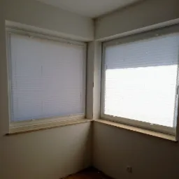 Dwa okna w rogu pokoju z zamontowanymi białymi plisami, widoczne prowadnice i sznurki. Parapety z beżowego kamienia, ściany w odcieniach bieli.