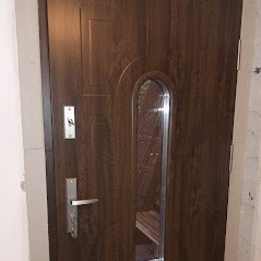 Brązowe drzwi wejściowe z pionowym, owalnym przeszkleniem i srebrną klamką, widoczne od wewnątrz, zamontowane w białej futrynie.