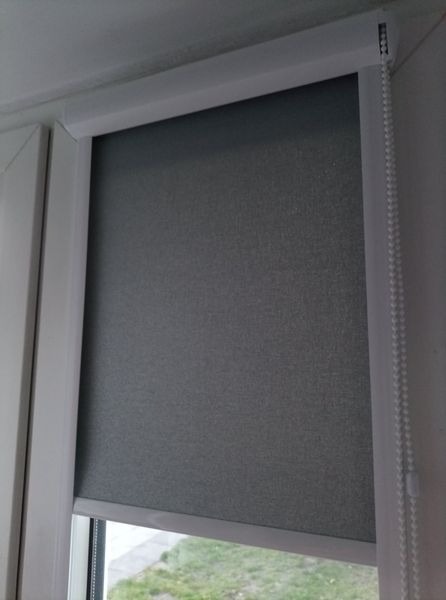 Szara roleta materiałowa zamontowana w białej ramie okiennej, widoczny mechanizm łańcuszkowy z boku, okno uchylone z widokiem na zieleń.