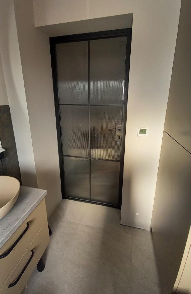 Szklane drzwi wewnętrzne z czarną ramą, podzielone na sześć segmentów, w nowoczesnej łazience z beżową umywalką i szarą podłogą. Widoczny termostat na ścianie.