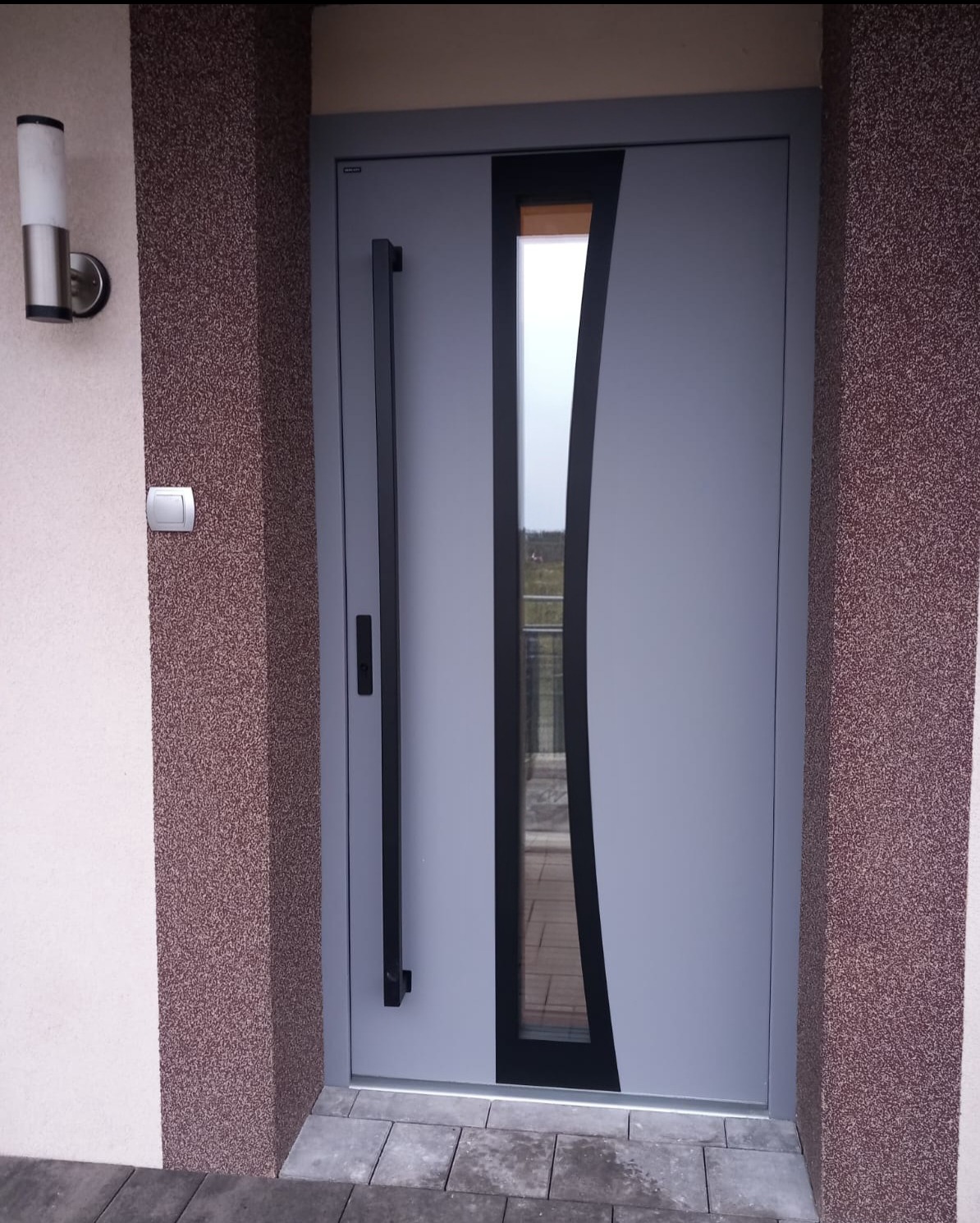 Szare drzwi zewnętrzne z długim, czarnym uchwytem i pionowym, zakrzywionym przeszkleniem, zamontowane w elewacji z brązowym tynkiem strukturalnym.
