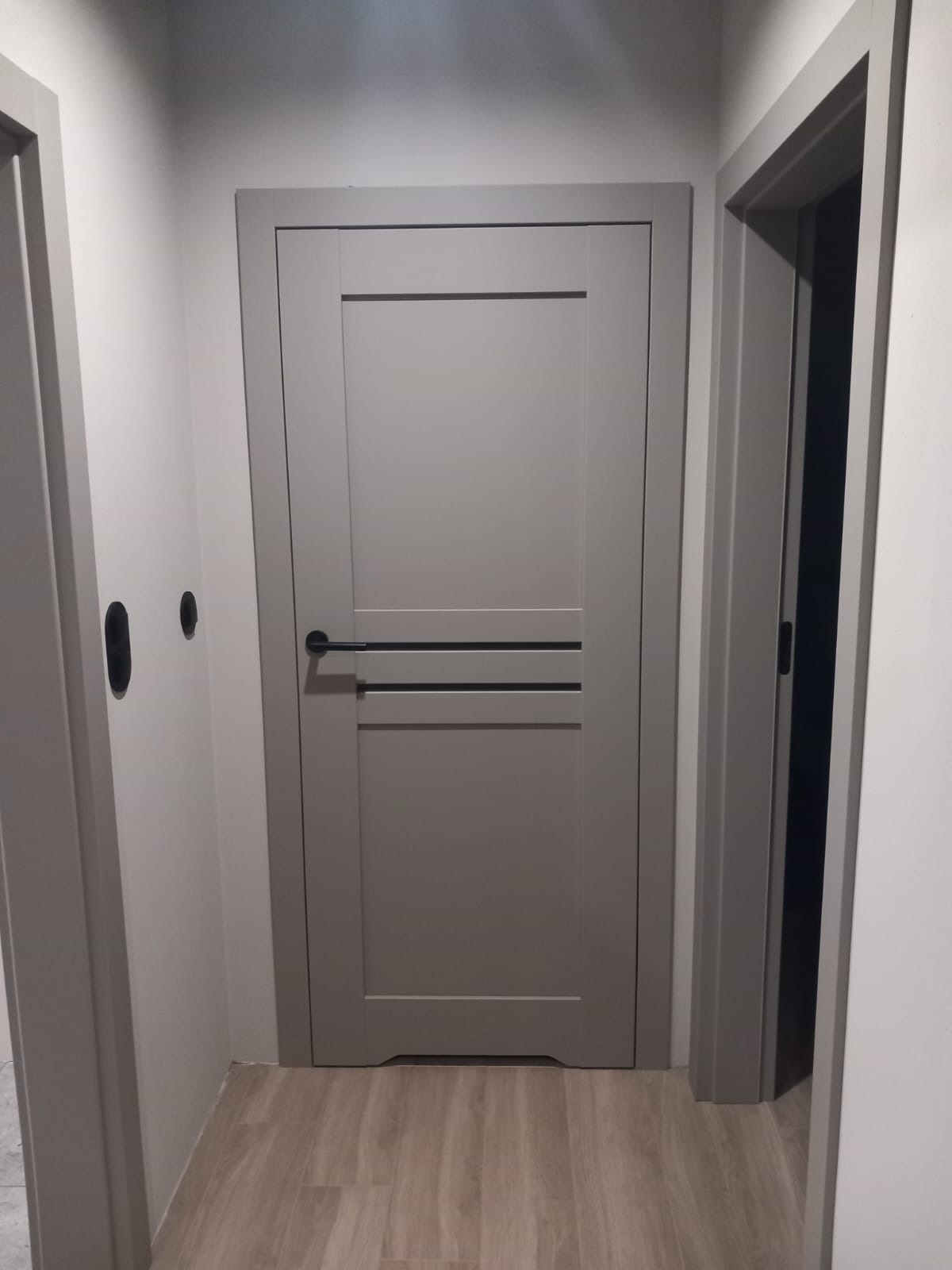 Szare drzwi wewnętrzne z czarną klamką i poziomymi zdobieniami, widoczne w korytarzu z podłogą imitującą drewno. Minimalistyczny design.