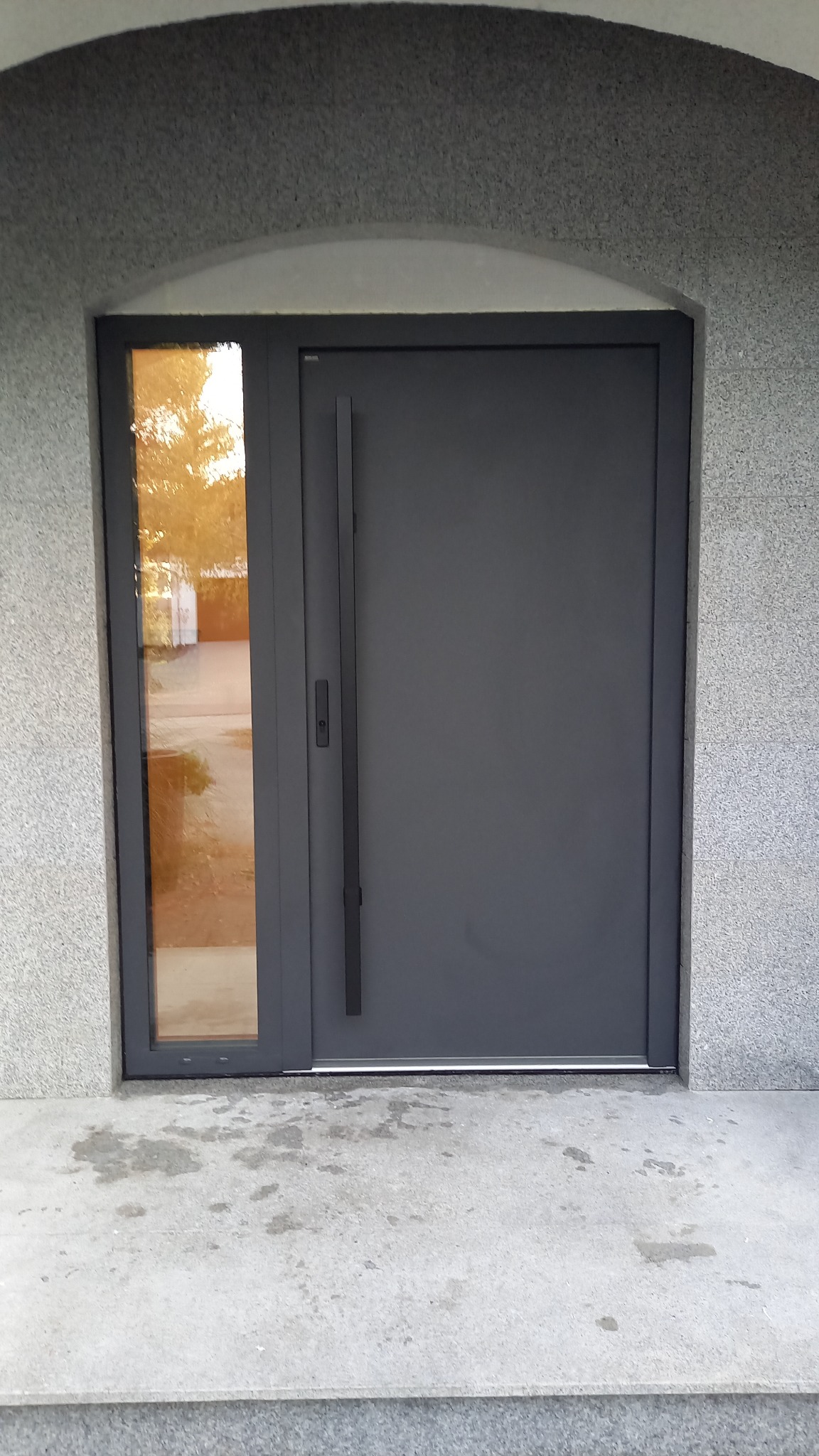 Grafitowe drzwi zewnętrzne z szybą boczną, długa klamka, osadzone w elewacji z szarego kamienia. Widoczny fragment schodów wejściowych.