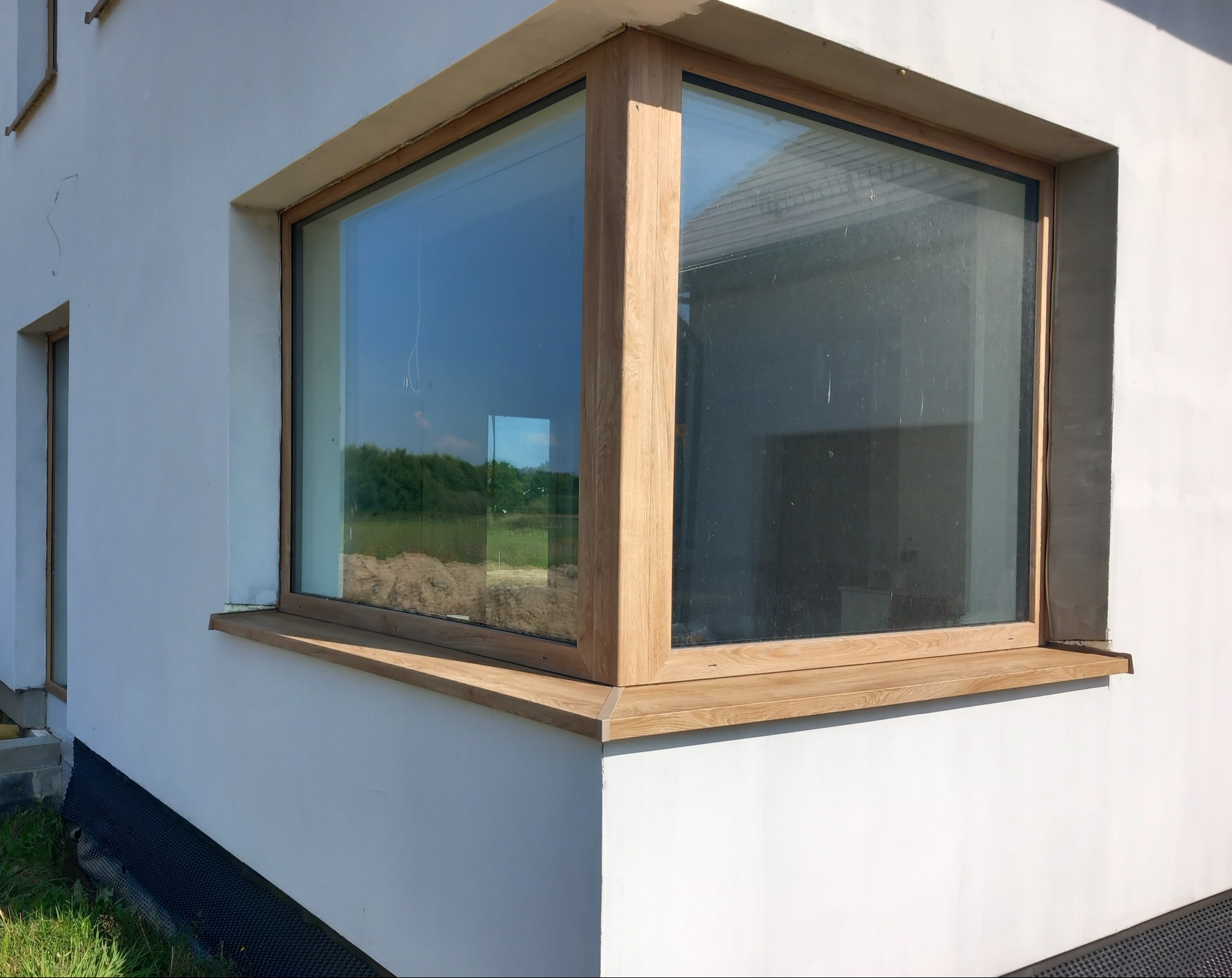 Narożne okno z drewnianą ramą i parapetem w białym budynku. Widok na zewnątrz przez szybę. Detal wykończenia elewacji i stolarki okiennej.