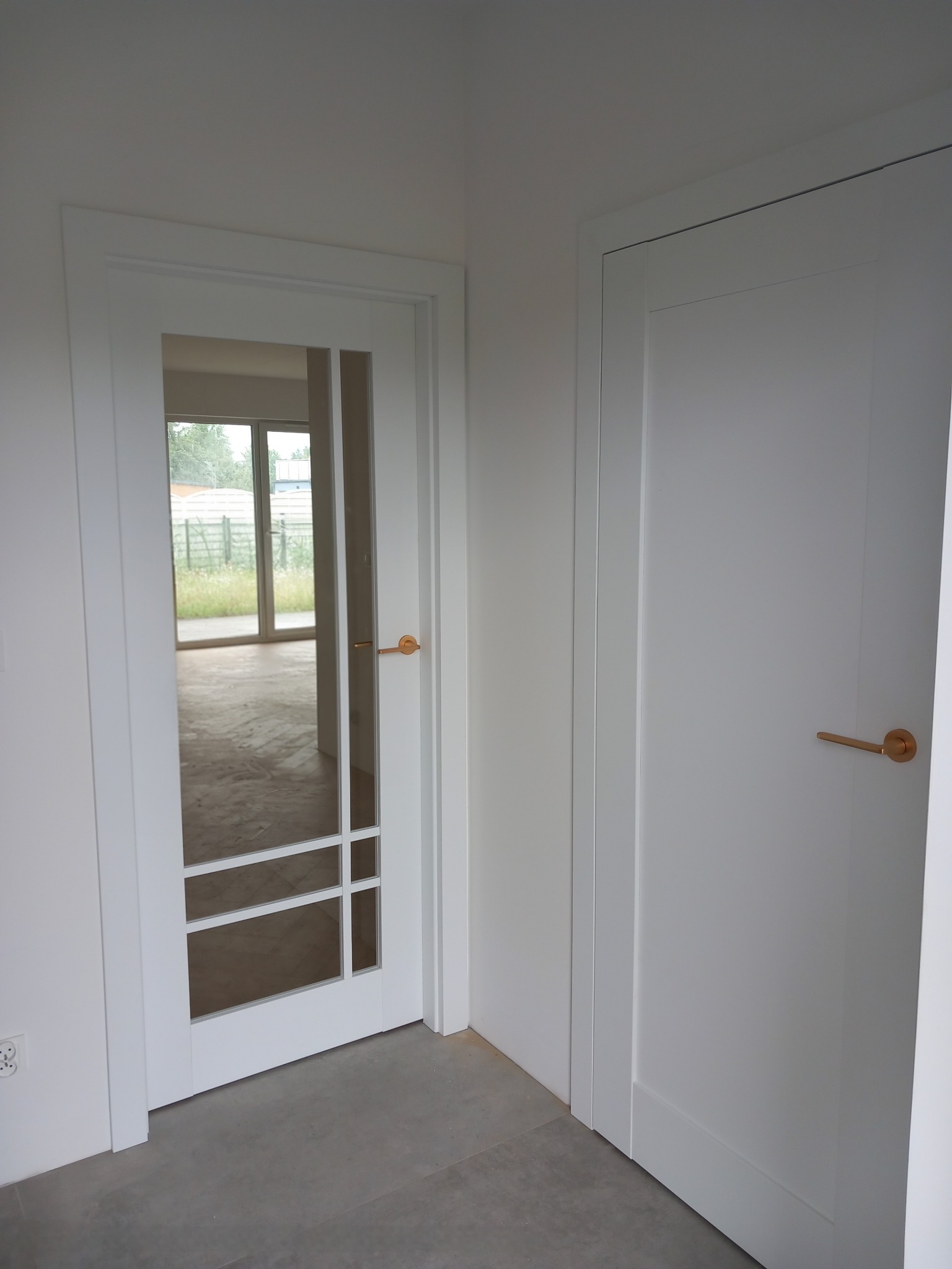 Białe drzwi wewnętrzne: skrzydło pełne i przeszklone z złotą klamką, widok na surowe wnętrze przez szybę. Minimalistyczny design, neutralna kolorystyka.