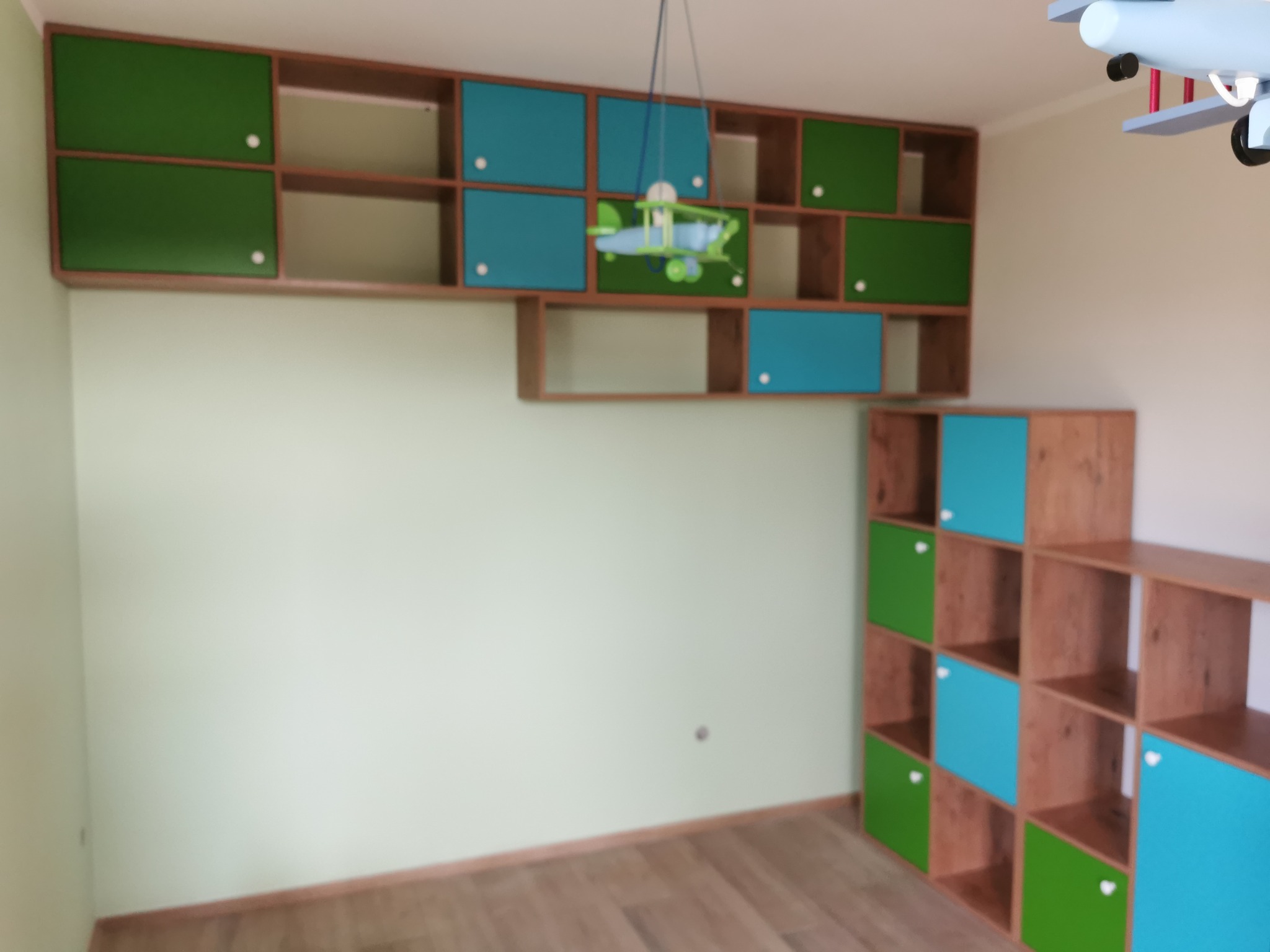 Narożne szafki ścienne i regał w pokoju dziecięcym, z zielonymi i niebieskimi frontami, zabawkowy samolot zawieszony na suficie, drewniana podłoga.