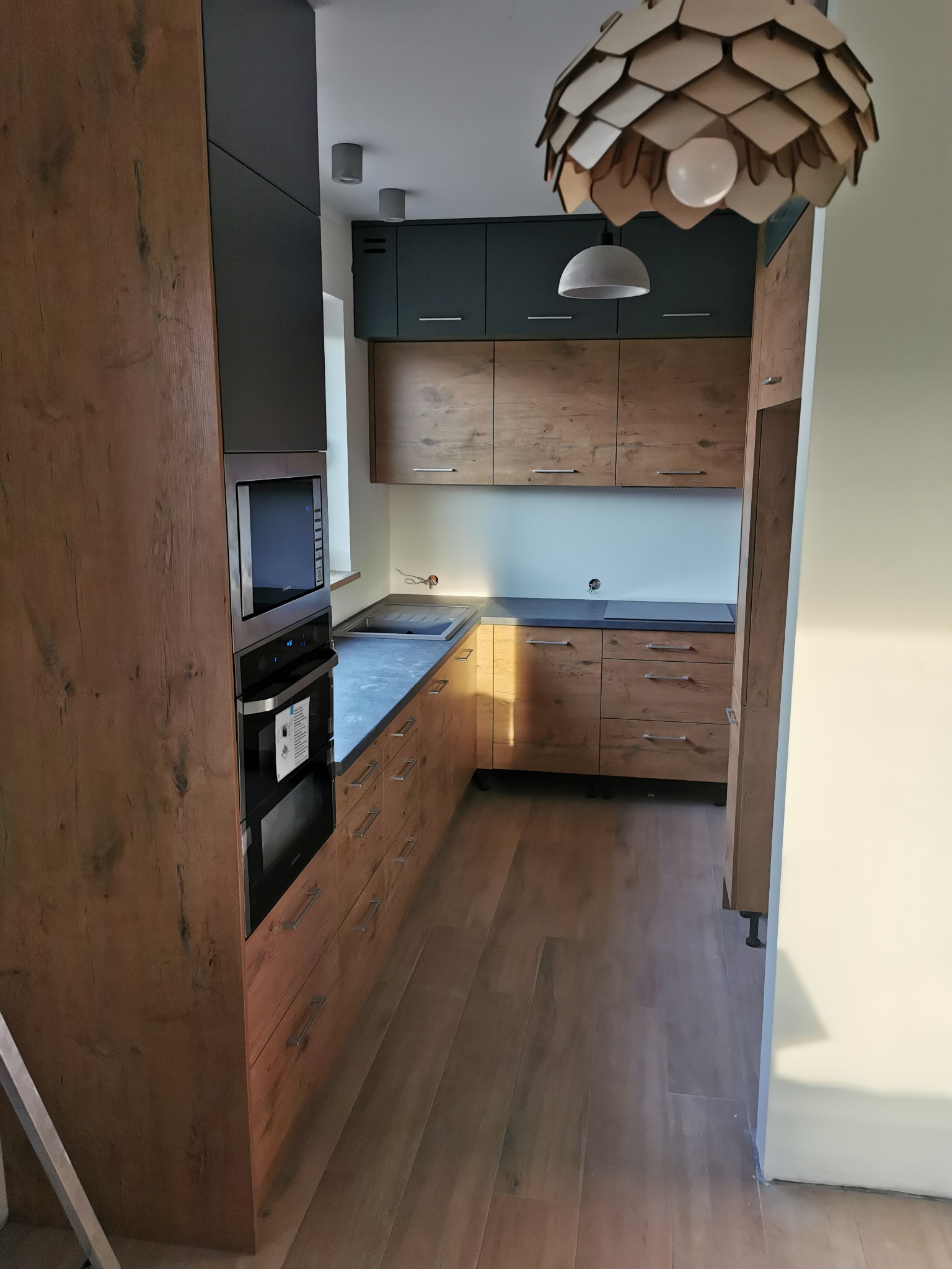 Nowoczesna kuchnia z drewnianymi frontami, ciemnoszarymi szafkami górnymi i blatem, oświetlona wiszącą lampą z drewnianych elementów, widok na całą długość pomieszczenia.