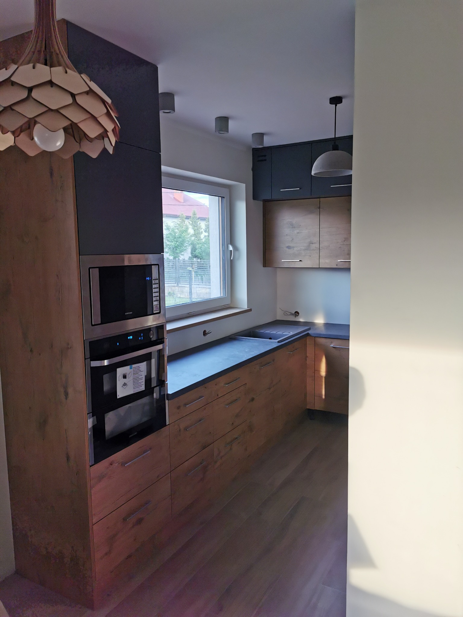 Nowoczesna kuchnia na wymiar z drewnianymi frontami i czarnymi akcentami w Warszawie, z wbudowanym piekarnikiem i mikrofalówką oraz designerską lampą wiszącą.