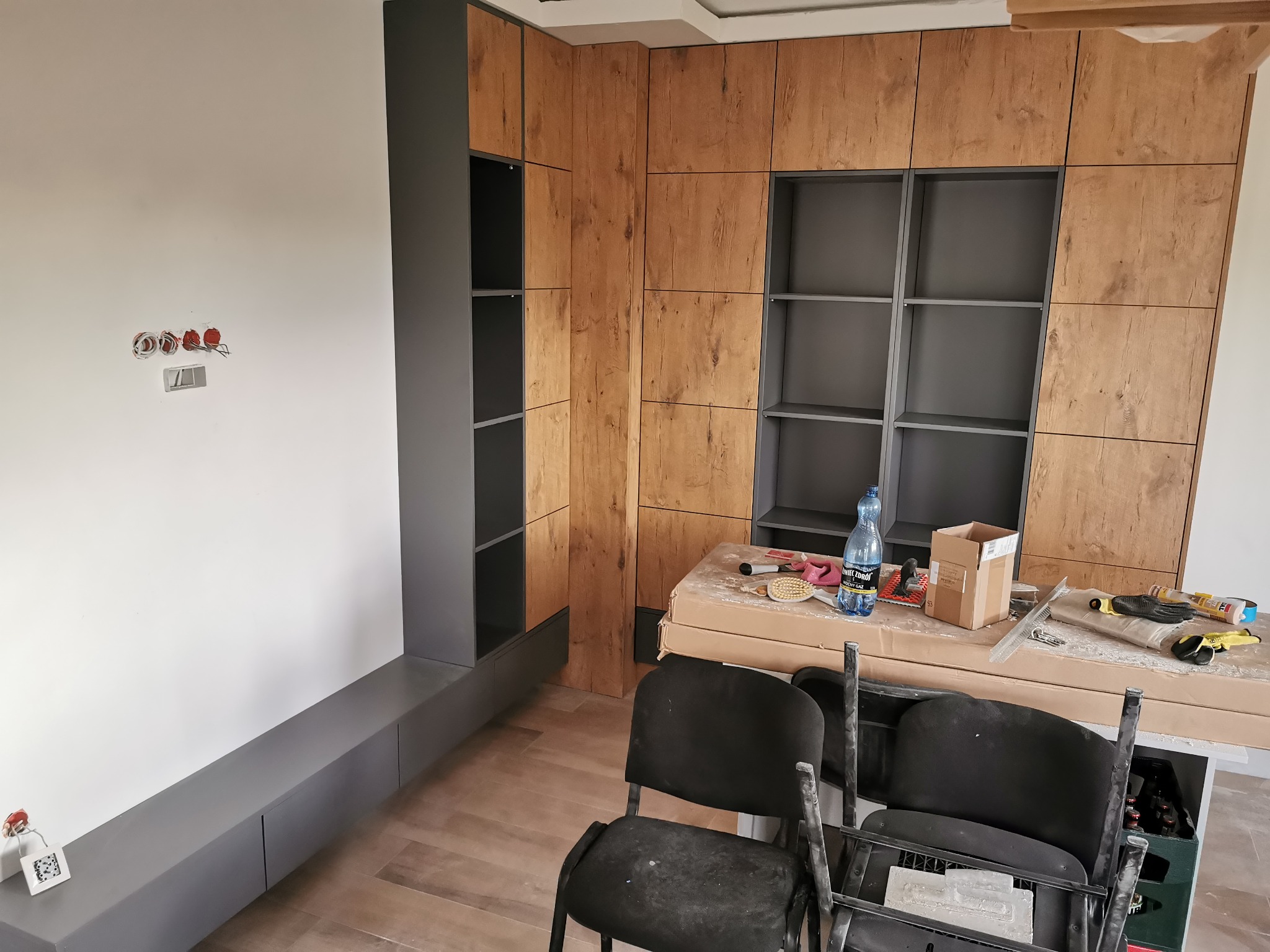 Montaż zabudowy meblowej z drewnopodobnymi frontami i szarymi elementami, widoczne narzędzia i materiały na stole roboczym, wnętrze w trakcie wykańczania.