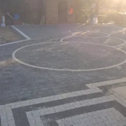 Ułożona nawierzchnia z kostki brukowej w odcieniach szarości z dekoracyjnymi elementami w kształcie okręgów i prostokątów, widok na budynek w tle w słoneczny dzień.