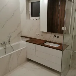 Nowoczesna łazienka z białymi marmurowymi płytkami, drewnianym blatem i białą szafką umywalkową, kabina prysznicowa ze szkła, minimalistyczna armatura.