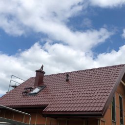 Konstrukcje drewniane i wykończenia Kacper Chmielewski - Czerwony dach z blachy trapezowej, komin i okno dachowe na tle błękitnego nieba z chmurami, widoczna konstrukcja domu szkieletowego z oknami.