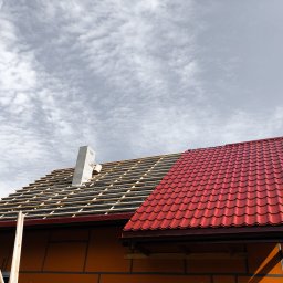 Konstrukcje drewniane i wykończenia Kacper Chmielewski - Częściowo pokryty dach z czerwonej blachodachówki, widoczne drewniane łaty i komin w trakcie budowy na tle pochmurnego nieba.