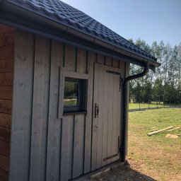 Konstrukcje drewniane i wykończenia Kacper Chmielewski - Drewniany budynek gospodarczy z oknem i drzwiami, pokryty szarą dachówką, z widocznym systemem rynnowym, na tle zielonego trawnika i drzew.