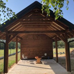 Konstrukcje drewniane i wykończenia Kacper Chmielewski - Drewniana altana z dachem dwuspadowym, widoczne krokwie i słupy nośne, obok leżą materiały budowlane, w tle zielony trawnik i drzewa.