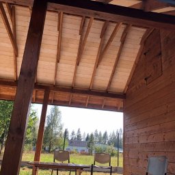 Konstrukcje drewniane i wykończenia Kacper Chmielewski - Wnętrze drewnianej konstrukcji z odsłoniętym drewnianym stropem i ścianami z desek, widok na zewnątrz przez otwartą przestrzeń, w tle zieleń i budynek.