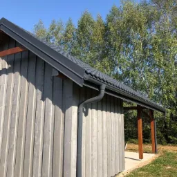 Szary, drewniany budynek z ciemnoszarą dachówką i systemem rynnowym, widok z zewnątrz na tle drzew i błękitnego nieba.