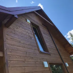 Brązowy dom z drewnianą elewacją w stylu skandynawskim, widok z dołu na fragment ściany z oknami i dachem z blachy trapezowej, na tle błękitnego nieba z chmurami i zieleni drzew.