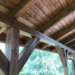 Drewniana konstrukcja altany ogrodowej z widocznym dachem z desek, słupami nośnymi i elementami ozdobnymi, pomalowana bejcą w odcieniach brązu i szarości, z zielenią w tle.