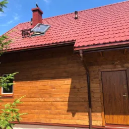 Wykończony dom szkieletowy z czerwoną dachówką, drewnianą elewacją, oknem dachowym i brązowymi rynnami, sfotografowany w słoneczny dzień z fragmentem iglastego drzewa w kadrze.