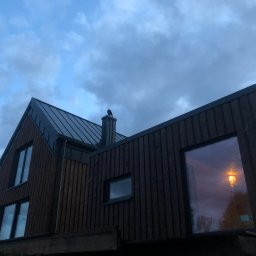 Konstrukcje drewniane i wykończenia Kacper Chmielewski - Nowoczesny dom z ciemną, pionową drewnianą elewacją i metalowym dachem, sfotografowany od dołu pod pochmurnym niebem. W dużym oknie odbija się ciepłe światło wnętrza, a na kominie siedzi ptak.
