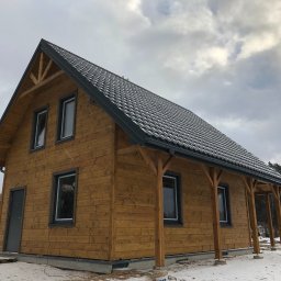 Konstrukcje drewniane i wykończenia Kacper Chmielewski - Nowoczesny dom z bali o jasnym, naturalnym wykończeniu ścian zewnętrznych i ciemnym, lekko ośnieżonym dachu dwuspadowym. Charakterystyczne drewniane wsporniki i zadaszony ganek. Otoczenie zimowe.