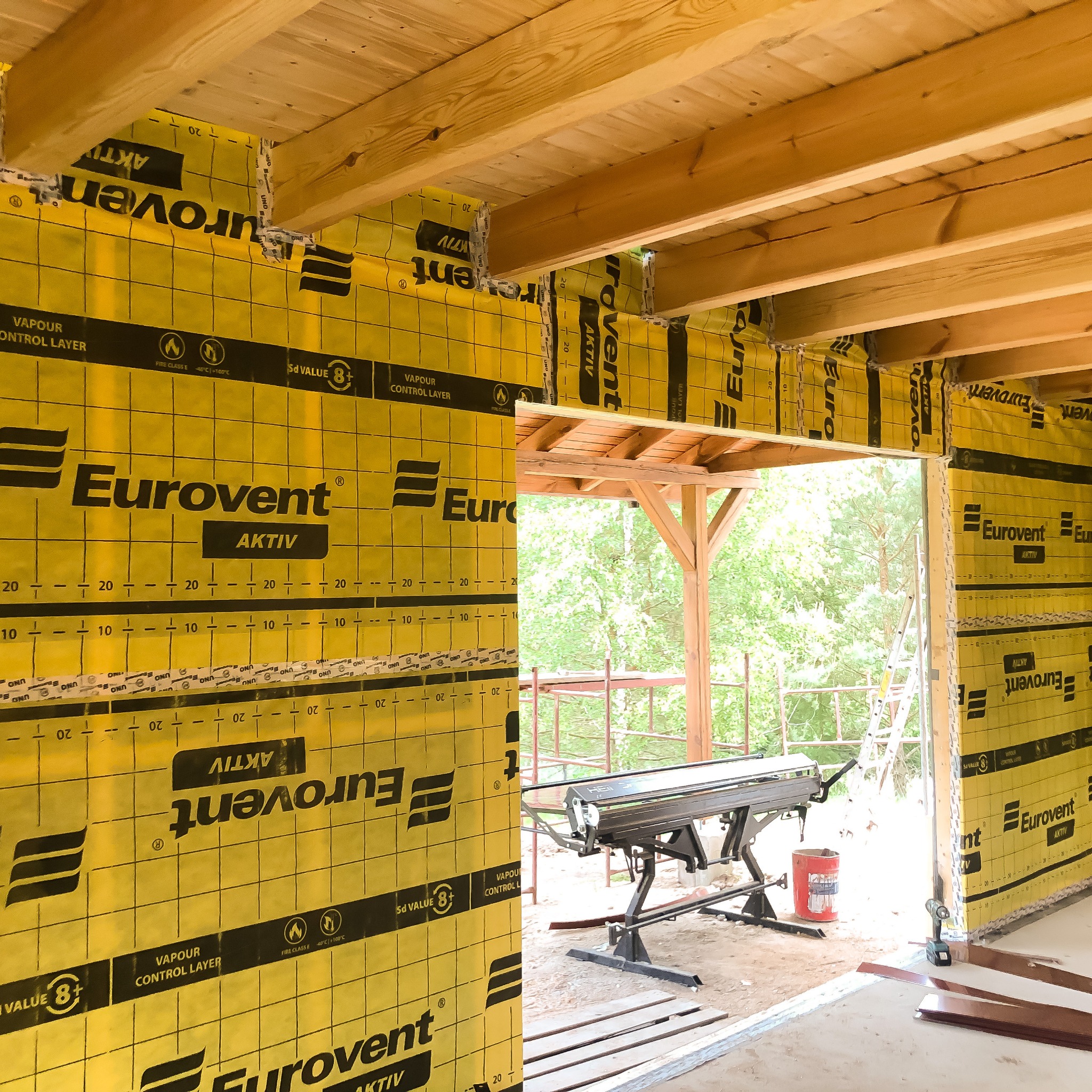 Wnętrze domu szkieletowego w trakcie budowy, widoczne drewniane belki stropowe i ściany pokryte żółtą folią paroizolacyjną Eurovent Aktiv z widocznymi oznaczeniami Sd Value 8+ i Vapour Control...