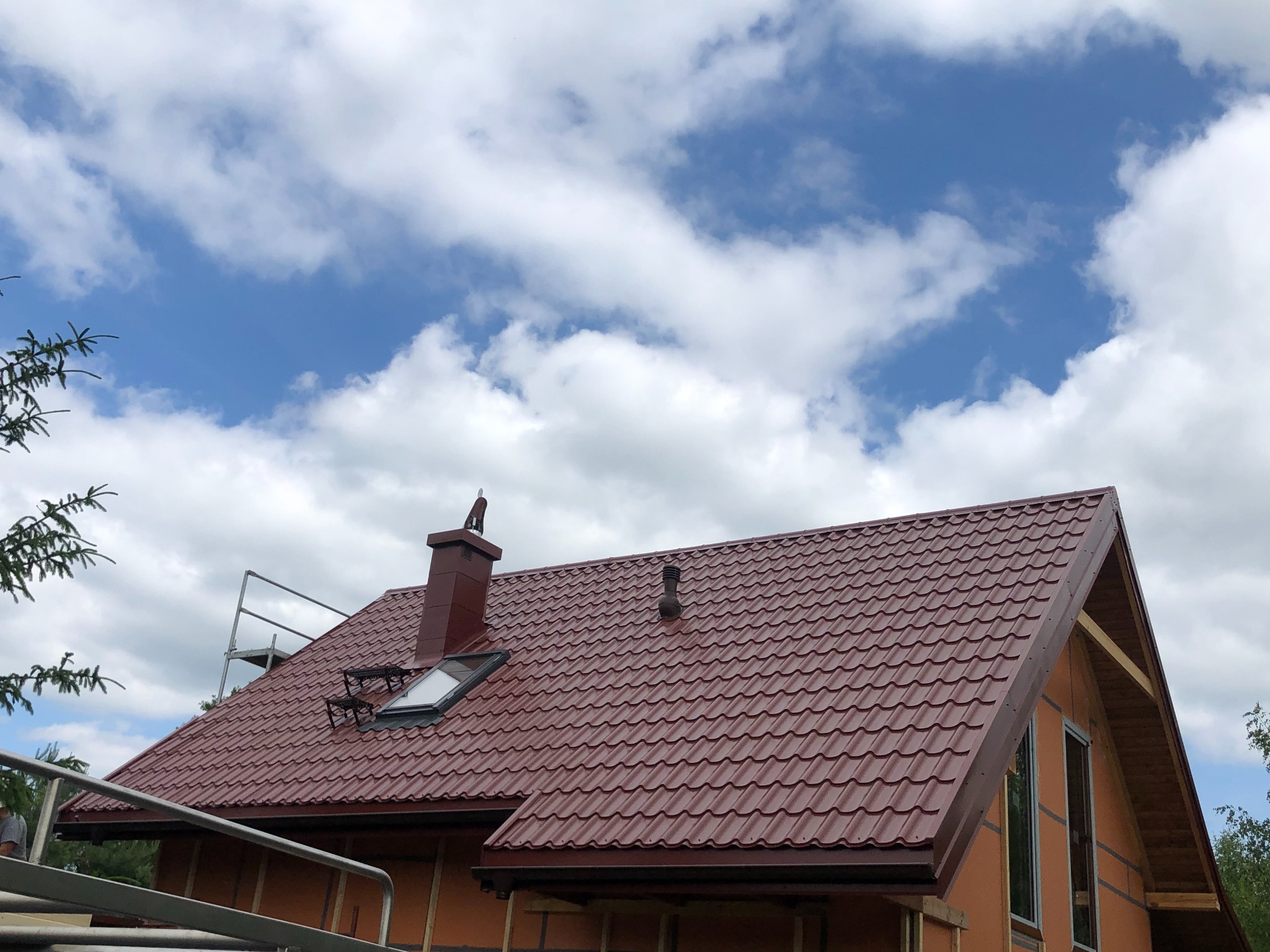 Czerwony dach z blachy trapezowej, komin i okno dachowe na tle błękitnego nieba z chmurami, widoczna konstrukcja domu szkieletowego z oknami.