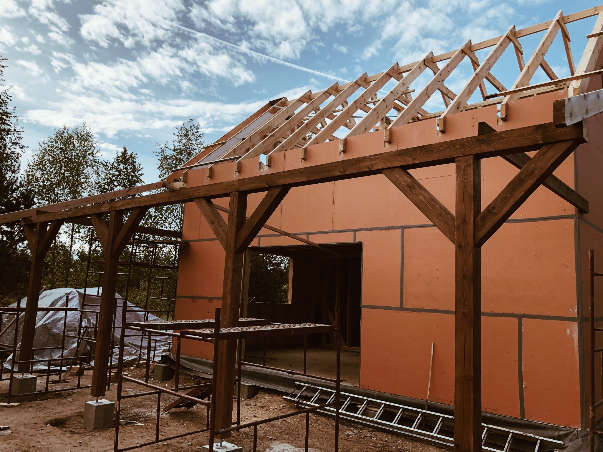 Budowa domu w technologii szkieletowej, widoczna konstrukcja drewniana ścian i dachu, częściowo osłonięta płytami, rusztowanie i drabina na pierwszym planie, błękitne niebo w tle.