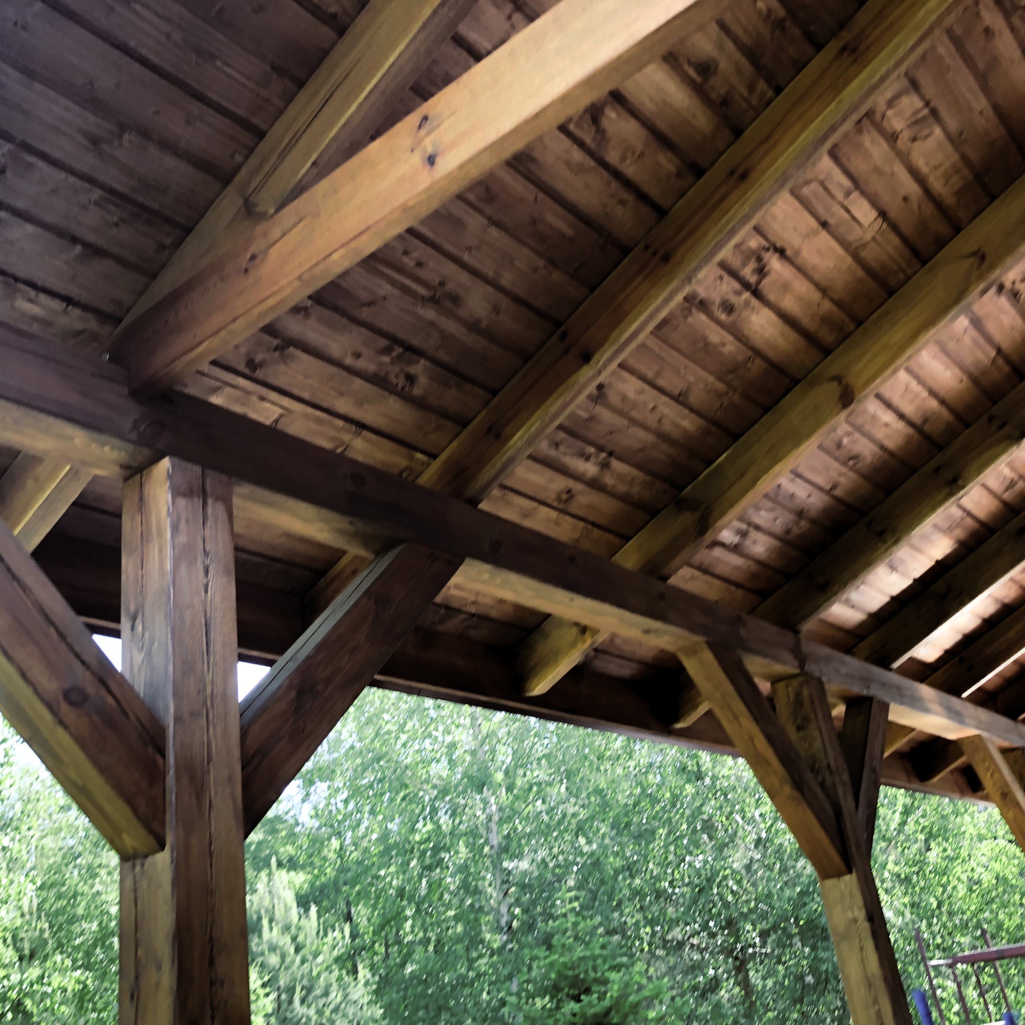 Drewniana konstrukcja z belek i desek, prawdopodobnie altana lub zadaszenie tarasu, widok od dołu, na zewnątrz widoczne drzewa.