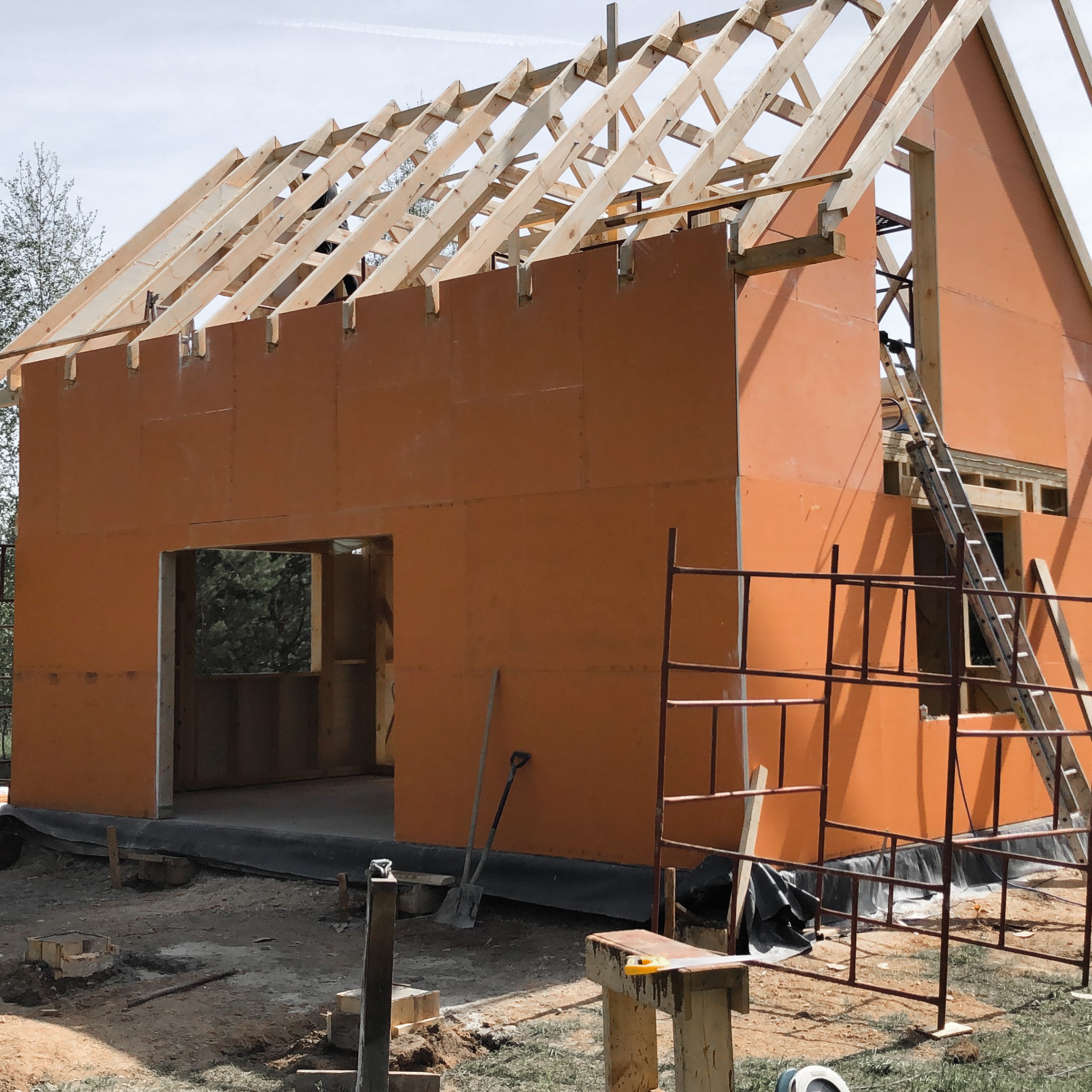 Wznoszenie ścian domu szkieletowego z widoczną konstrukcją dachu i rusztowaniem, na zewnątrz leżą narzędzia budowlane.