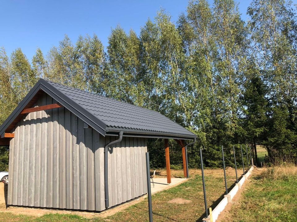 Niewielki budynek z szarą elewacją z desek i grafitowym dachem z blachodachówki, widok z zewnątrz na tle wysokich drzew.