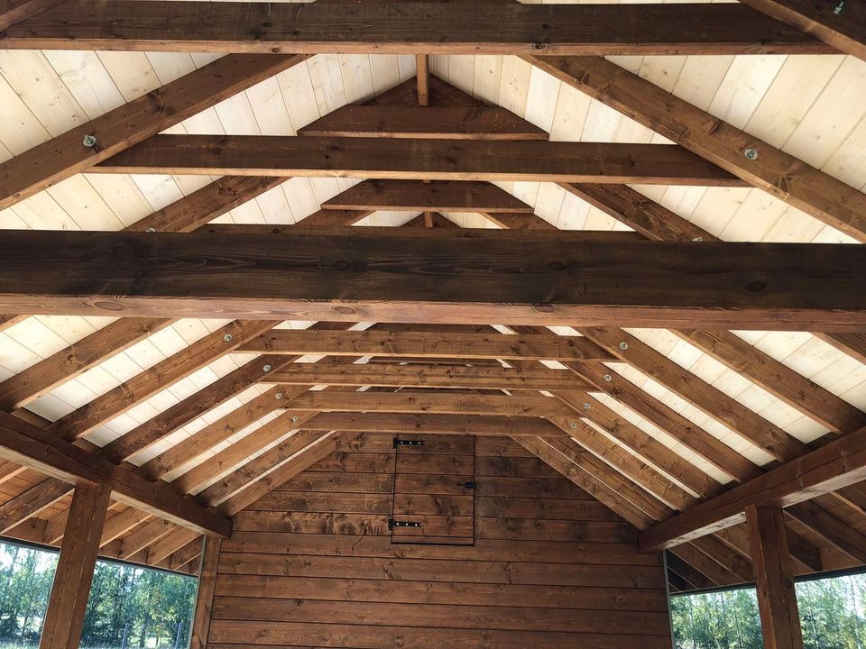 Wnętrze drewnianej konstrukcji z widocznymi belkami stropowymi i deskami, ciemne drewno ścian kontrastuje z jasnym wykończeniem dachu, widoczne okno z zielenią na zewnątrz.