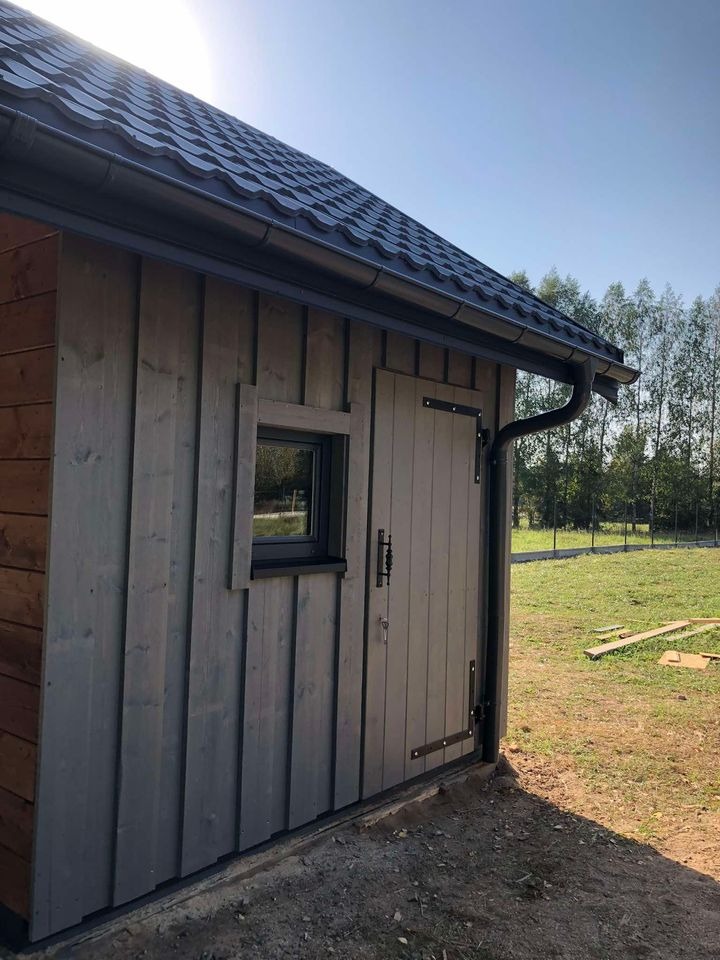 Drewniany budynek gospodarczy z oknem i drzwiami, pokryty szarą dachówką, z widocznym systemem rynnowym, na tle zielonego trawnika i drzew.