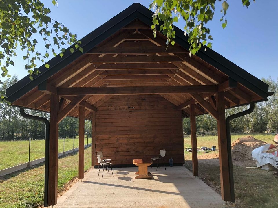 Drewniana altana z dachem dwuspadowym, widoczne krokwie i słupy nośne, obok leżą materiały budowlane, w tle zielony trawnik i drzewa.