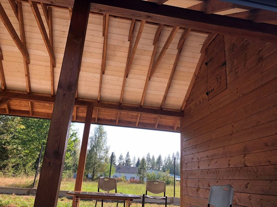 Wnętrze drewnianej konstrukcji z odsłoniętym drewnianym stropem i ścianami z desek, widok na zewnątrz przez otwartą przestrzeń, w tle zieleń i budynek.