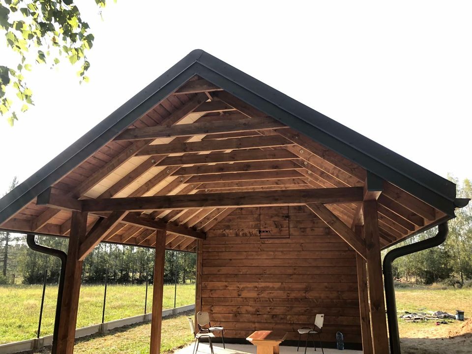 Drewniana altana z ciemnym dachem i rynnami, widok z zewnątrz na tle łąki i drzew, z dwoma krzesłami w środku.