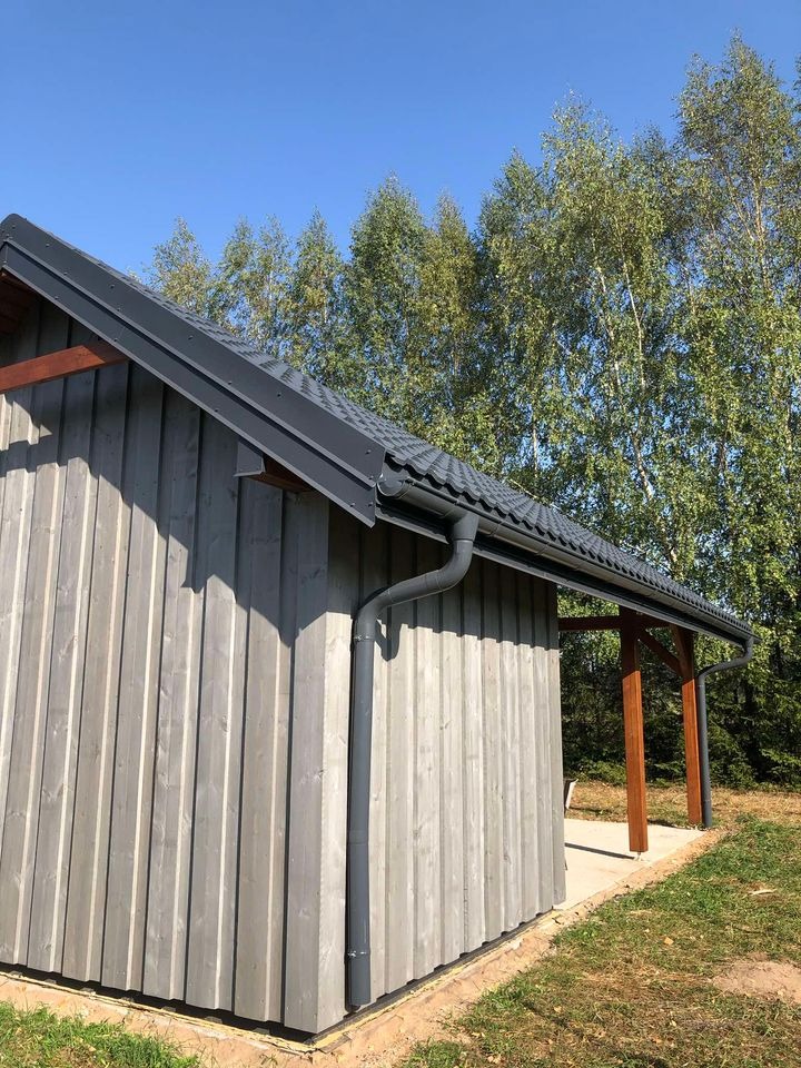 Szary, drewniany budynek z ciemnoszarą dachówką i systemem rynnowym, widok z zewnątrz na tle drzew i błękitnego nieba.
