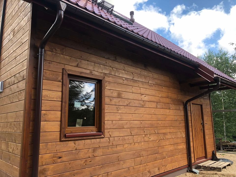 Gotowy dom szkieletowy z drewnianą elewacją, brązowymi rynnami i czerwoną dachówką, widok z boku na tle błękitnego nieba.