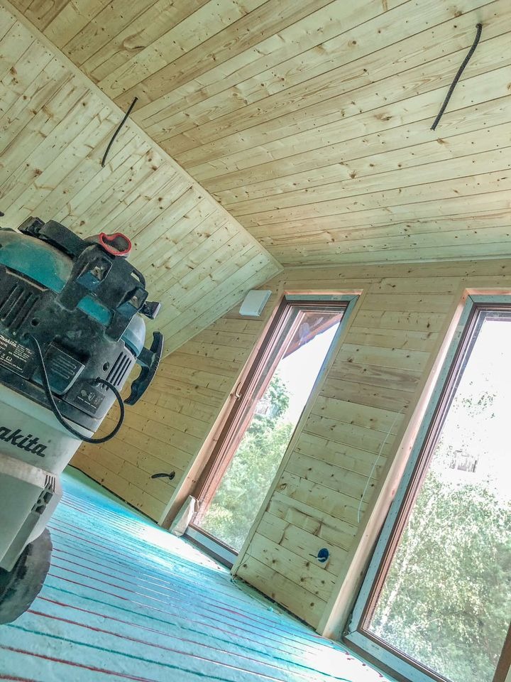 Wnętrze domu w trakcie budowy, widoczne drewniane ściany, skośny sufit z desek, okna dachowe oraz instalacja ogrzewania podłogowego i odkurzacz przemysłowy.