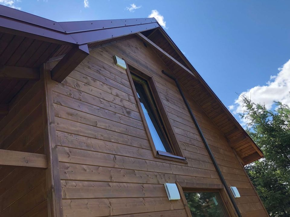 Brązowy dom z drewnianą elewacją w stylu skandynawskim, widok z dołu na fragment ściany z oknami i dachem z blachy trapezowej, na tle błękitnego nieba z chmurami i zieleni drzew.