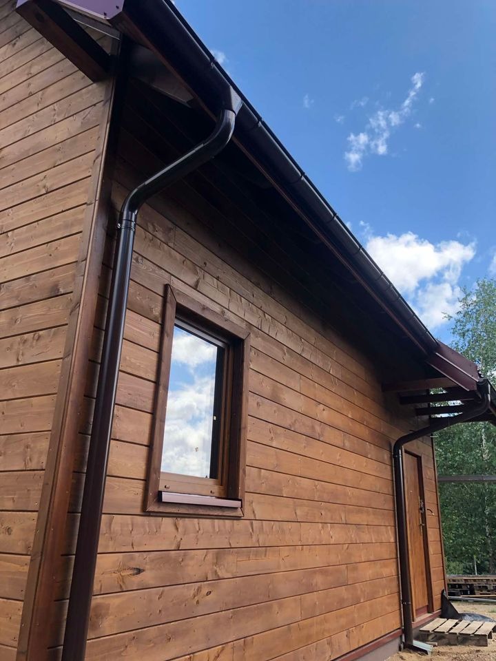 Brązowy dom z drewnianą elewacją, widoczne okno z odbiciem nieba i chmur, brązowe rynny i fragment dachu, jasne niebo w tle.