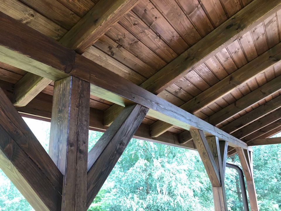 Drewniana konstrukcja altany ogrodowej z widocznym dachem z desek, słupami nośnymi i elementami ozdobnymi, pomalowana bejcą w odcieniach brązu i szarości, z zielenią w tle.