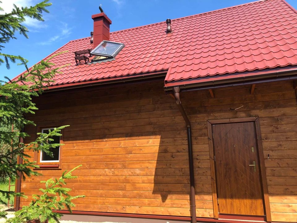 Wykończony dom szkieletowy z czerwoną dachówką, drewnianą elewacją, oknem dachowym i brązowymi rynnami, sfotografowany w słoneczny dzień z fragmentem iglastego drzewa w kadrze.